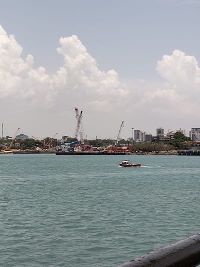 The image displays a coastal scene within Mkomani ward, Kenya, under a clear sky with scattered white cumulus clouds. The foreground consists of turquoise-green water with light surface ripples. A small, red and white motorboat is present in the mid-ground, traveling from right to left, leaving a visible wake. The distant shoreline reveals a working port area, characterized by numerous industrial cranes of various sizes, including at least two prominent lattice-boom cranes with red and white segments. Several large, dark-hulled vessels are moored along the shore. Behind the port facilities, a cityscape featuring multi-story buildings rises above green vegetation. A dark, out-of-focus railing or structure is visible at the bottom right corner of the frame. No individuals are distinctly identifiable.