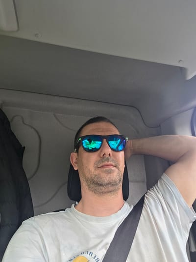 A male subject is depicted seated in the driver's position within a vehicle, specified as located in the city of Mid Suffolk, United Kingdom. The individual has short dark hair and facial stubble, and wears a light-colored t-shirt with a partial graphic. He is wearing black-framed sunglasses with highly reflective blue-green lenses. The reflections in these lenses clearly show a road scene with green verges and trees, consistent with forward travel. A dark-colored seatbelt is fastened diagonally across his chest. His left arm is bent, with his hand positioned behind his head. To the left of the subject, a black padded jacket is visible. The vehicle's interior features grey fabric and plastic panels, with a black headrest behind the subject. Small, light-colored protrusions are visible on the upper interior ceiling panels.