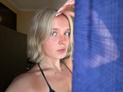 A young Caucasian woman with blonde hair and blue eyes is partially visible, leaning into the frame from the left. She is looking towards the camera with a slight smile and her hand is resting near her face, displaying long, decorated fingernails. She is wearing a black bikini top and a delicate necklace with small beads. A portion of a textured, deep blue fabric is draped over the left side of the image, obscuring part of her body and the background. The background appears to be an indoor setting with dark wooden furniture. The lighting suggests it could be daytime, possibly indoors near a window, or with artificial lighting. The mood seems relaxed and personal, possibly a selfie taken in a bedroom or dressing area. There is no visible text in the image.