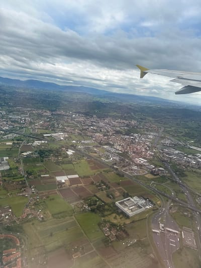 An aerial photograph, taken from an airplane window, captures a vast landscape under an overcast sky. The wing of the aircraft, featuring a yellow tip, is visible in the upper left corner. The sky is dominated by thick, grey clouds, with patches of lighter clouds and hints of blue sky.
The scene below reveals a mix of urban and rural environments, consistent with the area around Sesto Fiorentino, Italy. A dense urban sprawl characterized by numerous buildings and a complex network of roads covers a significant portion of the midground. A prominent multi-lane highway with an intricate interchange, including a large toll plaza and several ramps, dissects the landscape. Near the interchange, a large, modern building complex, possibly a commercial or industrial facility, stands out.
Beyond the urban areas, the landscape transitions into agricultural land, featuring numerous rectangular fields of varying shades of green and brown. Several white-roofed greenhouses are distinctly visible within these cultivated plots. In the distant background, a range of hills or mountains stretches across the horizon, appearing hazy beneath the cloudy sky. The photograph depicts a daytime scene, showcasing the blend of developed infrastructure and cultivated land from a unique aerial perspective. No text is visible in the image.