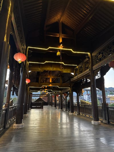 The image depicts an upward view of a traditional wooden structure, possibly a covered bridge or walkway, in Zhuoshui, China. The scene is illuminated with yellow LED strips, highlighting the intricate woodwork and design of the structure. Red lanterns are also present. The wooden walkway is visible on the right, with a view of the city beyond.