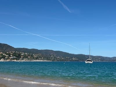 This image depicts a clear, sunny day at a beach in Le Lavandou, France. The foreground features a sandy beach with gentle waves lapping at the shore. The turquoise water extends to the horizon, where a line of green, tree-covered hills forms the backdrop. Along the coastline, numerous white sailboats are docked in a harbor, with many houses scattered across the hillside. In the middle ground, a white catamaran is anchored in the bay, its tall mast piercing the clear blue sky. Contrails from airplanes streak across the sky, suggesting air traffic above. The overall scene conveys a peaceful and idyllic coastal atmosphere.