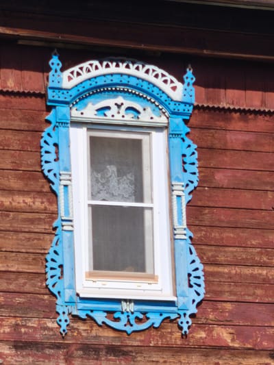 The image shows a close-up, eye-level view of a wooden window frame decorated with intricate blue and white carved wooden trim. The window itself is white-framed and appears to be made of glass with a white lace curtain visible inside. The window is set into a weathered wooden wall, painted a dark reddish-brown. The trim is ornate, featuring geometric patterns, stylized floral motifs, and decorative elements that extend from the sides and top of the window. The wood of the wall is aged, with visible grain and some signs of wear. The lighting suggests it is daytime, with sunlight casting shadows and highlighting the texture of the wood. The image focuses solely on the window and its immediate surroundings, giving no indication of the broader environment or location beyond the architectural style, which is common in rural Russian architecture.