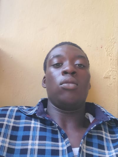 A close-up portrait features a young individual, likely male, with dark skin, looking directly at the camera. The person is wearing a blue, dark blue, and white plaid long-sleeved shirt, with the collar unbuttoned. The image is oriented at an angle, with the subject's head tilted towards the right. The background is a plain, light yellow-orange wall, showing some texture and minor imperfections, suggesting a simple indoor or sheltered outdoor setting. The lighting is soft and even, indicating daytime conditions. The person's expression is neutral to slightly contemplative, with open eyes. No other objects or specific details are visible beyond the individual and the wall. This portrait was taken in Atani, Nigeria. There is no visible text in the image.