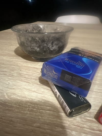A medium shot captures a wooden table with a glass bowl filled with a dark, granular substance. To the right of the bowl, a blue pack of Winston XS Compact 100's cigarettes rests on top of a black lighter, which has a metallic gold trim and a white striped pattern along its side. The scene is dimly lit, suggesting an indoor setting at night. The background is dark, with a faint blurry light visible in the upper left corner. The table surface exhibits a light wood grain pattern. There is no indication of the city of Krasnye Orly, Russia, or any people in this image.