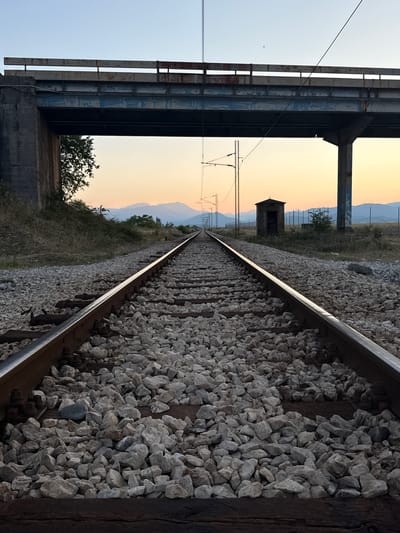 The image captures a railway track extending towards a distant horizon under a clear sky transitioning into sunset colors. A concrete bridge spans overhead, supported by pillars. To the right of the tracks, a small, weathered structure, possibly a signal box, stands near a fence. The surrounding landscape is dry and sparsely vegetated, with mountains visible in the background. The scene appears to be located in or near Mojanovići, Montenegro, given the typical terrain and infrastructure found in such areas. There are no people or animals visible in the image, and no discernible actions are taking place.