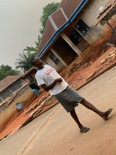 A man wearing a white t-shirt and gray shorts stands on a dirt road. He has sandals on his feet. Behind him is a building, partially constructed, with exposed brick and visible windows. Green foliage surrounds the building, suggesting a natural setting. The area appears rural or developing, with a mix of modern construction and more traditional building styles.