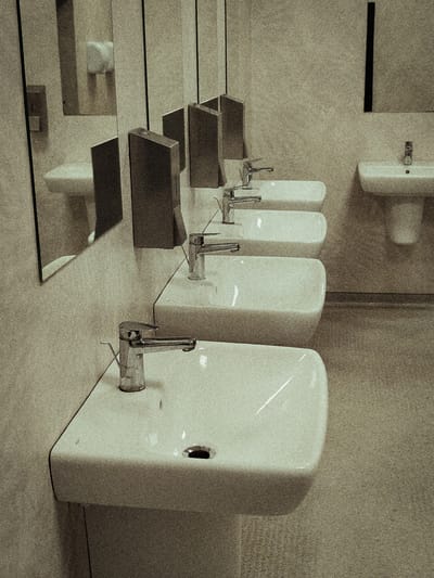 The image depicts a row of white ceramic sinks with chrome faucets in a public restroom. The sinks are modern and rectangular, and the faucets are of a lever-operated design. The environment appears clean and well-maintained, with tiled walls and a speckled floor. Mirrors are present, reflecting the sinks and the restroom's lighting. The scene is static, with no people visible. The overall impression is one of a functional and minimalist space. This restroom is located in the city of Łomna-Las, Poland.