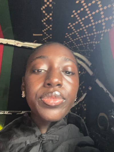 The image shows a close-up, selfie-style portrait of a young Black woman. She has dark skin, dark eyes, and full lips that appear to be covered in lip gloss. She is wearing small, sparkling earrings. The background is dark with a pattern of small, light-colored squares arranged in a diamond-like motif, and vertical stripes of green and red are visible on the left side. She is wearing a black garment that is tied at the neck with a knot. The image appears to have been taken indoors. The location is identified as Karmajiji, Nigeria.
