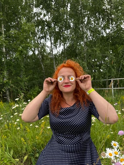 A Caucasian woman with vibrant red, curly hair and red lipstick is holding two daisies in front of her eyes, resembling sunglasses. She is wearing a dark blue dress with white polka dots and a yellow wristband. The woman is outdoors in a grassy field filled with white and yellow wildflowers, with a backdrop of green trees. The image appears to be taken in Privolzhsky, Russia, suggested by the yellow wristband with Cyrillic text.