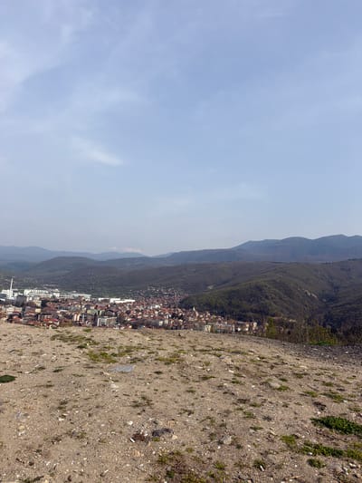 The image shows a landscape view in Sveti Konstantin, Bulgaria. The perspective is from an elevated, rocky, dirt area overlooking the town, which is nestled between a mountain range and what appears to be a slightly hazy sky. The town is densely built with a mix of low-rise buildings, and a few taller structures. The mountains are covered with dense vegetation. The ground in the foreground consists of dirt, rocks, and sparse patches of grass.