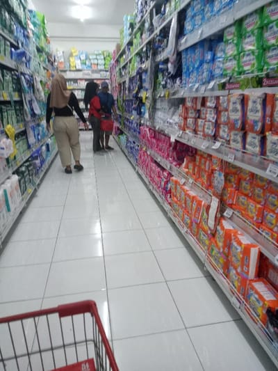 This image depicts an interior view of a supermarket aisle in Nganjuk, Indonesia. The perspective is from behind a red-handled shopping cart, partially visible in the bottom-left corner. The aisle features a clean, white tiled floor with visible grout lines and reflections from overhead fluorescent lighting. Two rows of tall, metal-shelved displays, stocked with various consumer goods, extend into the background on both sides.
Three individuals are present in the aisle. In the mid-ground, walking away from the camera, is a person wearing a beige hijab, a black long-sleeved top, and light brown trousers, with black shoes. Further down the aisle, two other individuals are standing next to the right-hand shelves. One wears a red shirt and holds a red shopping basket, while the other wears a blue shirt and possibly a dark cap, appearing to browse the products.
The shelves on the left contain a variety of packaged items, primarily in white, light blue, and green packaging, likely personal care or household products. The shelves on the right are densely packed with products, predominantly sanitary pads or diapers, distinguished by their bright pink, blue, orange, and white packaging. Some packages bear text in Indonesian, such as "2X CEGAH." The lighting is bright and even throughout the retail space.