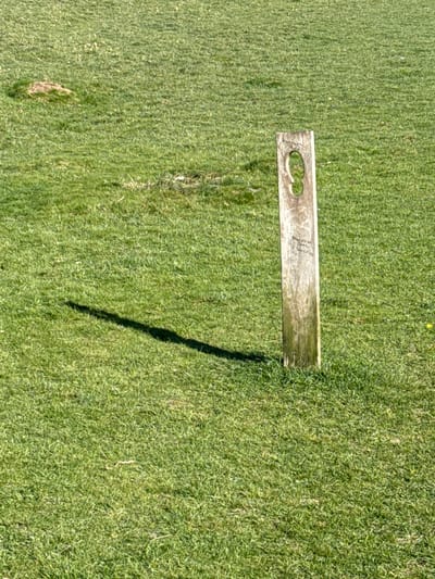 The image shows a weathered, rectangular wooden object lying on a field of green grass. The object has at least one rounded cutout feature. A shadow extends from the object onto the grass, suggesting the presence of sunlight. The image appears to be taken from a top-down perspective. The location is specified to be in Dacorum, United Kingdom, but there are no visual cues in the image to confirm this.