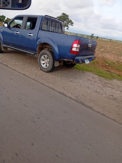 The media file shows the rear passenger side of a blue Ford pickup truck parked on the side of a road. The truck has a standard cab and a black roll bar in the bed. The truck's tires are dirty, suggesting it has been driven on unpaved roads. The road itself is gravelly and appears to be in a rural area. In the background, there is a dry, brown field with scattered trees and hills in the distance. The sky is overcast with clouds.
The scene appears to be set during the daytime, though the exact time is not discernible. The weather seems to be cloudy and possibly warm, typical of a dry season. The location cues, such as the dry vegetation and the type of truck, suggest a rural or semi-arid environment. There is no visible text in the image. The overall impression is one of a functional vehicle in a natural, undeveloped setting, possibly in Jauro Sent, Nigeria, as per the location context. The perspective suggests the photo was taken from another vehicle moving alongside the truck.