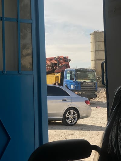 An outdoor scene features a white car parked near construction vehicles. A blue Scania truck is visible, along with a crane and a concrete mixer. The vehicles are parked on a sandy surface. The shot is framed through what appears to be a blue window or doorway with a window pane design. The composition includes part of a car seat and interior roof lining in the upper part of the image.