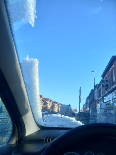 The image depicts a street scene in Noyelles-sous-Lens, France, viewed from inside a vehicle on a bright, clear day following snowfall. Patches of snow and ice adhere to the vehicle's windshield, notably along the left A-pillar and across the lower central glass area. The vehicle's dark steering wheel and a portion of the dashboard are visible in the foreground.
The street extends into the distance under a cloudless blue sky. On the left, a continuous row of two-story residential buildings with light-colored facades and reddish-brown tiled roofs lines the road. On the right, two-story commercial or mixed-use buildings with darker brick facades are present. A prominent sign for "MARKET CITY" is affixed to the upper right side of one building. Below this, reflective material is propped against a storefront. A dark, vertical street light pole is also visible on the right.
Several vehicles are present on the street: a white van is in the mid-ground, followed by a dark sedan. Another white car is visible further down the street on the left. Residual patches of snow or slush are visible on the road surface and along the curbs, consistent with the snow on the windshield. No individuals are discernible within the frame.