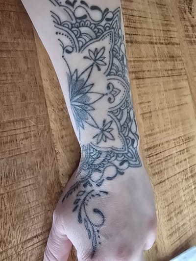 The image shows a close-up, overhead view of a person's left forearm and hand, which is adorned with a black ink tattoo. The tattoo is intricate and features floral and mandala-like patterns extending from the wrist up the forearm. The skin is fair. The forearm and hand are resting on a wooden surface with a prominent grain pattern. The background is blurred, indicating a shallow depth of field. There is no discernible information to suggest a location in Karlstadt, Germany.
