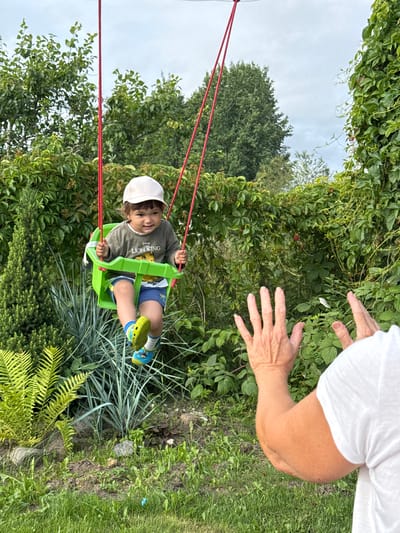 In Sitno, Russia, a young child wearing a white cap, a dark green long-sleeved shirt with "The Lion King" graphics, and blue shorts is seated in a green baby swing. The child is smiling and looking towards the camera. The swing is suspended by red ropes from an unseen overhead support. In the foreground, a person's arms and hands are visible, reaching out towards the child as if to catch them or wave goodbye. The background consists of lush green foliage, including trees, bushes, and vines, under a cloudy sky. The setting appears to be a garden or backyard.