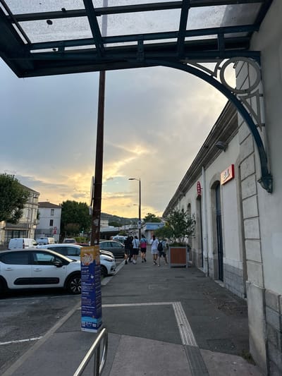 The image shows a street scene in Alès, France, likely near a train station given the presence of a covered walkway with a glass roof and a pole with advertising. The sun is setting, casting a warm glow on the sky and illuminating the clouds. In the foreground, a white car is parked on the left, and a metal railing is visible. A tall, cylindrical advertising pillar stands on the sidewalk, displaying event information. A group of people are walking away from the camera on the sidewalk, heading towards a building with shops. One building has a red "RELAY" sign and a red "TABAC" sign. Trees and other buildings are visible in the background, along with more parked cars and streetlights. The overall atmosphere is that of a late afternoon or early evening in a small city.