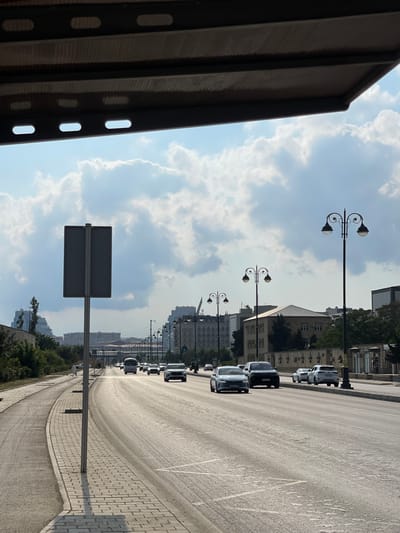 The media file depicts a street scene in Khatay Raion, Azerbaijan. The image is oriented vertically, with the sky occupying the upper portion and the street curving away into the distance. On the left side of the frame, a dark, corrugated structure partially obscures the view. The sky is partly cloudy, with large, cumulus clouds scattered against a pale blue backdrop.
A paved street runs diagonally through the scene, bordered by a sidewalk made of interlocking bricks. Several cars are parked or driving along the street, appearing as dark shapes from this perspective. Streetlights with ornate, traditional designs are visible along the roadside, interspersed with modern buildings in the background. These buildings are predominantly light-colored and have a block-like architectural style, suggesting an urban environment. Green trees are also visible, adding a touch of nature to the cityscape.
A prominent feature in the foreground is a large, gray, rectangular sign mounted on a pole, facing away from the viewer. The sign appears to be a traffic or informational sign, but its details are not discernible. The sunlight suggests it is daytime, likely late morning or early afternoon, given the angle of the shadows. The overall atmosphere is that of a quiet, ordinary day in a city. No people are clearly visible in the image. There is no discernible text in the image.