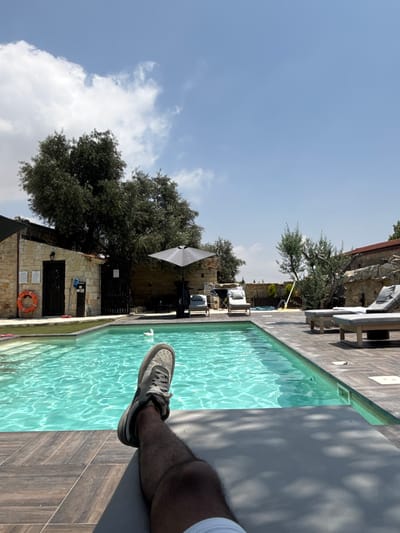 The image depicts a person's legs and feet relaxing on a lounge chair by a swimming pool. The pool is a bright turquoise color and surrounded by a wooden deck. In the background, there is a stone building, lush green trees, and a clear blue sky with some clouds. The scene appears to be a tranquil vacation setting. The location is identified as Pachna, Cyprus.