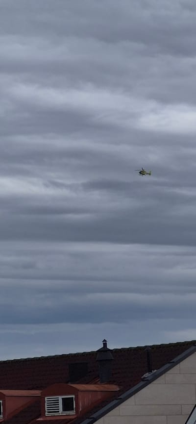 A small, yellow helicopter is seen in flight against a cloudy sky. The helicopter appears to be a light model, with visible rotor blades and a tail rotor. It is positioned in the upper left quadrant of the image, isolated against the vast expanse of clouds.

To the right of the helicopter, the side of a building is visible, indicating an urban or suburban setting. The building has a dark red, tiled roof with a steeply pitched gable. A dormer window with white bars is prominent on the upper part of the gable. The building's exterior appears to be constructed from brick and some lighter colored material, possibly stone or siding, in the upper section. A dark, angular fixture, possibly a ventilation cap or decorative element, sits on the roof near the peak of the gable.

The sky is filled with an overcast of grey clouds, suggesting a cloudy or overcast day. The lighting is diffused, typical of such weather. The time of day is not precisely discernible but the general light levels suggest it is daytime. The location context of Lillehammer, Norway, implies the scene is set in this Scandinavian city. No text is visible in the image. The overall mood conveyed is one of everyday activity, with the helicopter in transit and the building representing a residential or commercial structure.