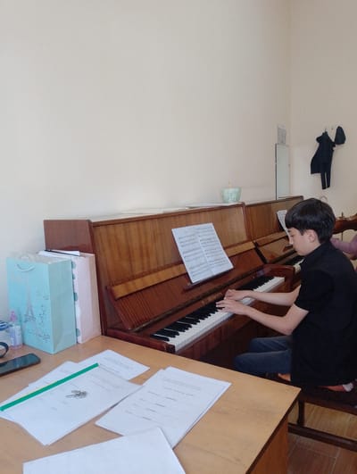 In a room with neutral-colored walls, a young person with dark hair and a black shirt is playing a piano. Sheet music is open on the piano, and the young person's hands are on the keys. In the background, a coat is hanging on a hook on the wall. To the left of the piano, there are several gift bags and a phone on a desk. The room appears to be a music studio or practice room. The specific location of Nor Hachin, Armenia, cannot be determined from the visual information provided in the image.