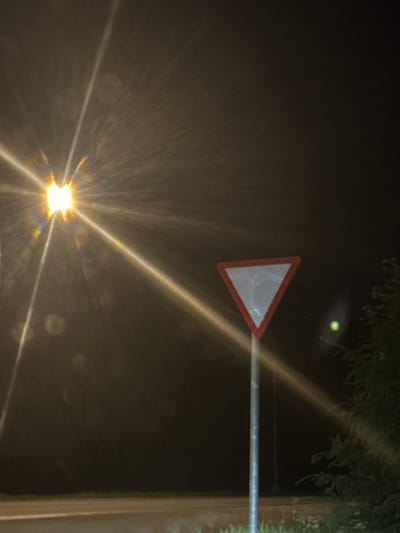An overhead street light casts a strong glare with visible rays and rainbow-like flares across the dark, hazy sky of Sof'ino, Russia. In the middle ground, a triangular yield sign with a red border and white interior is mounted on a pole. To the right, the silhouette of trees is visible against the dark background. The foreground shows a dimly lit paved surface, likely a road. A small, green light is visible in the mid-right portion of the image.