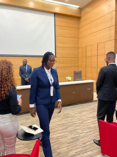 The image depicts an indoor scene from what appears to be a formal conference, meeting, or presentation in Luanda, Angola. The primary subject is a woman with dark dreadlocks and glasses, dressed professionally in a navy blue pantsuit over a white shirt. She is standing upright, looking towards the left side of the frame, and wears a blue lanyard with an ID badge that prominently features the word "AFRICA" and a blue and white logo, suggesting participation in an African forum or event.
Behind her, another man in a dark suit stands facing away, partially visible behind a light-colored table or podium adorned with a small white floral arrangement. To the left, a large white projection screen is mounted on the wall, indicating that a presentation or visual content is part of the event. A third man in a dark suit is visible in the distant background, further to the left, behind the main standing woman.
In the foreground, the back of a person's head with curly hair is visible, holding a smartphone. A small black stand with a laptop or tablet rests near this person. The venue features light-colored wooden paneling on the walls and a textured, patterned carpeted floor. Red chair backs are discernible on the right side of the frame, hinting at an audience area. The overall setting is professional and formal, illuminated by indoor lighting.