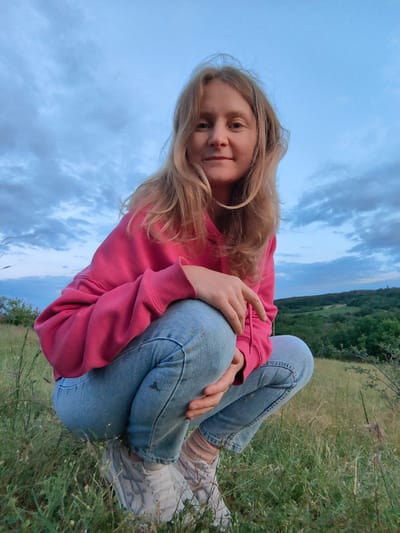 A young woman with blonde hair is crouching in a grassy field with a rolling, tree-covered landscape in the background. She is wearing a pink hooded sweatshirt and light blue jeans. The sky above is cloudy and appears to be either early morning or late evening. The overall mood is casual and relaxed. The image is taken from a low angle, with the woman looking directly at the camera.