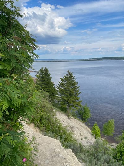 This media file captures a scenic landscape view from a high vantage point overlooking a wide river or large body of water, possibly near Пятый километр, Russia. The perspective looks down onto the water and across to the distant horizon.

In the foreground, dense green foliage, including shrubs with compound leaves and a distinct pink flower, frames the lower right portion of the image. The immediate ground slopes steeply, revealing a rocky and sandy cliff face. This bank is dotted with various green bushes and several dark green evergreen trees, primarily conifers. Lighter green deciduous trees grow closer to the water's edge at the base of the cliff.

The expansive body of water, with subtle ripples on its surface, dominates the midground, appearing dark blue and grey. In the distance, a long, low-lying bridge or elevated structure spans the water, connecting to a distant bank covered with a strip of dark green forest. The sky above is a mix of bright blue and scattered white to grey clouds, some wispy and others more substantial. The scene is bathed in natural daylight, indicating daytime, likely late morning or afternoon.

The image conveys a sense of natural beauty and tranquility. No people or visible text are present.