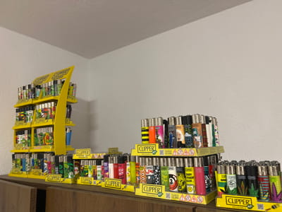 The image displays a collection of "Clipper" brand lighters arranged on multiple yellow display stands. A large, multi-tiered yellow display, prominently featuring the "CLIPPER" logo and "REUSABLE" text, occupies the left side of the frame. To its right, several smaller, single-tier yellow trays, also containing "Clipper" lighters and branded similarly, are linearly arranged. All display stands are placed on a wooden surface, likely a counter or shelf, which extends across the lower portion of the image. The lighters exhibit a wide variety of colorful designs, including abstract patterns, cartoon characters, pop culture motifs, and prominent cannabis leaf imagery. The background consists of a plain, light-colored wall. No individuals, actions, or specific interactions are visible. The scene is static and indoors. The image provides no discernible information regarding its specific location within the city of Mosomishte, Bulgaria.