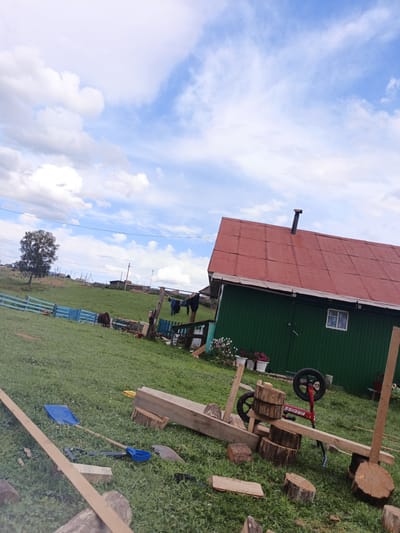The image depicts an outdoor scene in Islambaevo, Russia, likely a rural area. A green building with a red tin roof and a chimney is visible on the right. In front of the building, a makeshift structure appears to be under construction or assembly, utilizing logs and wooden planks. A small red bicycle with the word "KRCISS" on its frame is partially incorporated into this structure.
The foreground is a grassy area with various wooden logs and planks scattered around. Tools, including a blue shovel and another shovel, are also present on the grass, suggesting ongoing work. A blue fence is visible in the mid-ground, and behind it, a dark-colored animal, possibly a horse or cow, is grazing in a green field. A lone tree stands to the left of the animal. Further in the background, several rustic structures and fences indicate a village or settlement. The sky is partly cloudy with scattered white clouds against a blue backdrop. Clothes are hanging on a line in the mid-ground, near the green building.