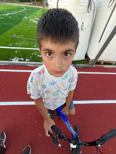 A young boy with dark hair and brown eyes stands on a red track, holding the handlebars of a blue bicycle. He is wearing a white t-shirt with colorful cartoon-like graphics and blue shorts. The track has white lines marking lanes. In the background, to the left, is a green artificial turf field with white markings, possibly a sports field. To the right, behind the boy, are large white sacks, possibly for storage or construction. The scene appears to be outdoors, in a recreational or industrial area. The image is a close-up shot, focusing on the boy and his immediate surroundings.