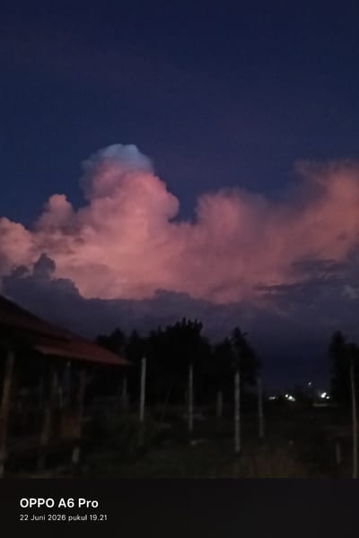 This is a vertical, low-light photograph of a dramatic sky over a rural landscape at dusk. The sky is a deep, dark blue, with large, puffy cumulus clouds that are illuminated from below by the setting sun, giving them a vibrant pink and light blue hue. Silhouetted against the colorful clouds are dark green trees and the roofline of a building with a reddish-brown roof. Several vertical poles are visible in the foreground and mid-ground. In the distance, small, blurry lights suggest a settlement or houses.
The time of day is indicated by the date and time stamp at the bottom of the image: "22 Juni 2026 pukul 19.21," which translates to June 22, 2026, at 7:21 PM. This time, combined with the colors in the sky, suggests sunset or twilight. The weather appears to be clear enough for a sunset, with dramatic cloud formations. The setting is likely a rural area in Blangkutacoh, Indonesia, given the context.
Visible text at the bottom of the image reads "OPPO A6 Pro," identifying the device used to take the photo. The overall mood evoked by the image is one of natural beauty and tranquility, with the vibrant colors of the sunset creating a striking contrast against the darkening sky and silhouetted landscape.
