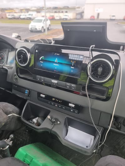 The image shows the interior of a vehicle, likely a van or truck, with a focus on the dashboard and center console. The dashboard features a large touchscreen display showing icons for "Phone," "Navigation," and "Radio." To the left of the screen is a vent and part of the instrument cluster, including a speedometer. Below the touchscreen are various buttons and controls, including climate control settings displaying "22°C" and icons for A/C, recirculation, and defrost. There are also storage compartments, one of which contains a set of keys. A green toolbox is visible on the floor of the vehicle. The exterior of the vehicle is partially visible through the windshield, showing a parking lot with several other vehicles, including white SUVs and vans, and some buildings in the background. The sky appears overcast. Based on the specific brand names visible on the items in the storage compartment (such as "GRIÓTGARDAR"), the location is identified as Njarðvík, Iceland. The scene appears to be a parked vehicle in an outdoor setting.