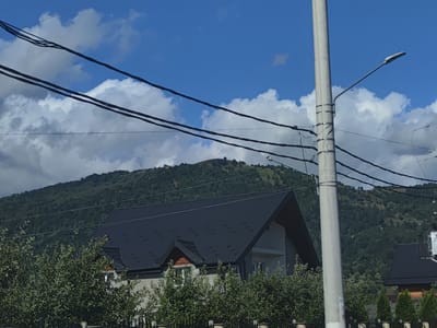 The image captures a view of a residential area in Petrova, Romania, under a bright blue sky with scattered white clouds. In the foreground, several dark power lines stretch across the frame. A tall, light-colored utility pole with a street lamp is positioned on the right side. Behind these, a dark-tiled roof of a house is prominent, with some trees and shrubbery partially obscuring it. Further in the background, a lush green, tree-covered mountain rises, its slopes dotted with more trees. The overall scene depicts a somewhat rural setting within a city.