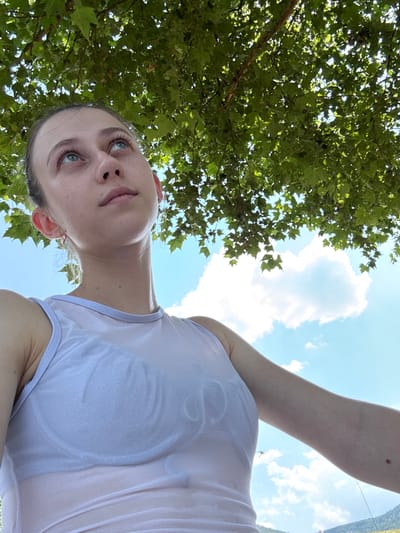 The image captures a young woman outdoors, positioned in Bürgstadt, Germany. She is looking upwards, with her face tilted towards the sky, which is visible through the canopy of green leaves from a tree overhead. The woman is wearing a white, sheer tank top over a white bra. Her arm is extended to her right, and a mole is visible on her forearm. The background consists of a blue sky with scattered white clouds and a hint of distant green hills. The lighting suggests it is daytime, likely a sunny day.