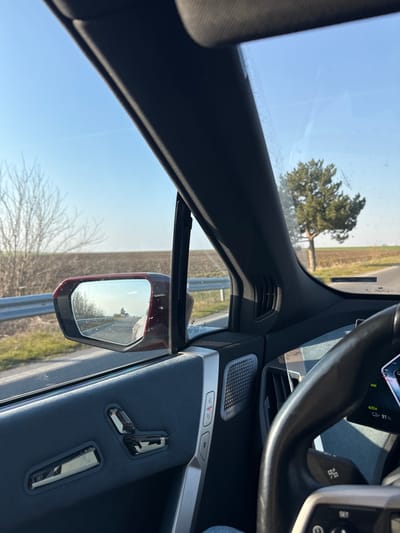 The scene depicts a view from the interior of a moving vehicle, looking forward and to the left through the passenger window and partially through the windshield. The vehicle appears to be a modern BMW, possibly an iX model, identifiable by the distinctive steering wheel design, the door panel with integrated electric seat controls (three position, one recline), a silver trim element, and a speaker grille bearing "Bowers & Wilkins" branding. A digital display, partially visible behind the steering wheel, shows "AUTO" and "91%", likely indicating an automatic driving mode and 91% battery charge. Dust or smudges are present on the inner surface of the vehicle's windows.
Outside the vehicle, a rural landscape is visible under a clear, bright blue sky with faint, linear contrails. To the immediate left, a red side mirror reflects the road extending behind the car, showing a distant pine tree. Adjacent to the road, a silver guardrail runs parallel, bordering a grassy verge. Beyond the verge are expansive, barren agricultural fields, light brown in color, stretching towards the horizon. A cluster of bare deciduous trees stands on the left side of the road further in the distance. To the right, through the windshield, a single, prominent pine tree with dense green foliage stands beside the paved road surface. The road curves slightly to the right in the distance. The environment suggests a daytime setting, possibly late autumn or early spring due to the bare trees and field conditions.
This rural road is located near Zlati Voyvoda, Bulgaria.