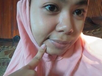 The media file is a close-up, eye-level, medium shot of a young person. The subject is wearing a pale pink hijab that covers their head and chest. Only the face and part of the torso are visible. The subject has a light brown complexion and dark brown eyes, which are looking directly at the camera. Their mouth is slightly parted, revealing their lips. The subject's right index finger is raised and pointing towards their chin, with the fingernail visible. The background consists of wooden vertical planks on the left, and what appears to be a patterned sofa or upholstery on the right, suggesting an indoor setting. The lighting is somewhat bright, with highlights visible on the subject's forehead and cheek, indicating it might be daytime or a well-lit indoor space. There is no visible text in the media file. The scene appears to be a casual, personal moment captured in Labissa, Indonesia.