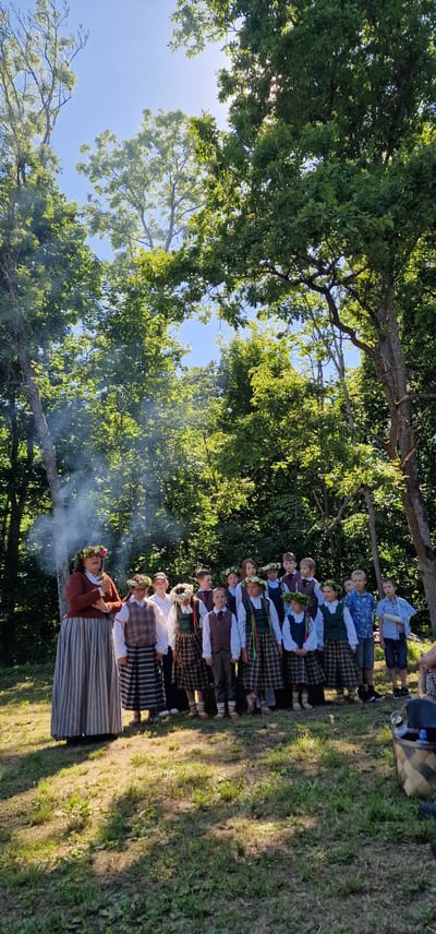 An outdoor gathering is taking place in a sunlit forest clearing, likely in Liepupe, Latvia. A woman, dressed in a long, striped skirt and a rust-colored top, stands at the left of the frame, wearing a floral crown. She appears to be addressing a group of children and young people, all of whom are also adorned with floral crowns. The children are dressed in traditional attire, with boys wearing vests over white shirts and checkered skirts or pants, and girls in similar skirts and vests. Some children are also wearing green ribbon accents. Behind the group, a faint plume of white smoke rises, possibly from a nearby fire or incense. The environment is characterized by lush green trees and dappled sunlight, creating a vibrant and natural setting.