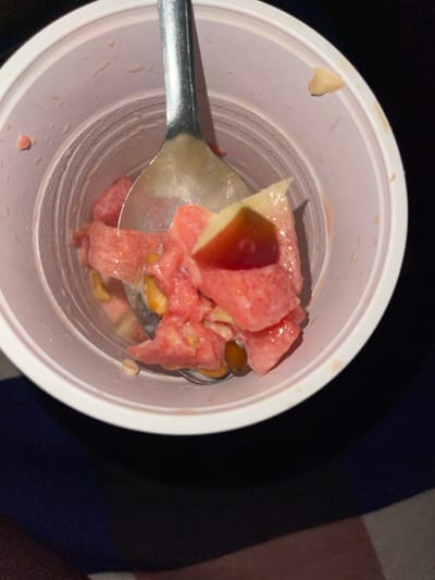 A top-down forensic view shows a light pink plastic cup containing a mix of diced fruit and nuts, located in Mape, Nigeria. The primary contents include numerous irregularly cut pieces of red-pink fruit, consistent with watermelon, and smaller, paler yellow fruit chunks, possibly apple or pear. Several ovoid, yellowish-brown items, resembling roasted peanuts, are interspersed within the mixture. A silver-colored metal spoon is partially submerged among the contents, indicating ongoing or recent consumption. The inner walls of the cup exhibit pink residue and moisture droplets. The immediate background is a dark, indistinct surface. No individuals are visible in the scene.