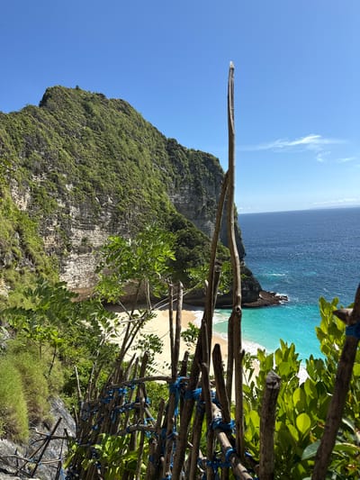 A landscape photograph taken in Bunga Mekar, Indonesia, shows a cliff overlooking a beach and ocean. The cliff face is covered with greenery. A stick fence is in the foreground on the right side. The ocean is blue and turquoise near the beach. The sky is blue with a few clouds. A long stick is pointing upward from the left.
