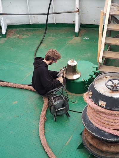 The image depicts a man working on the deck of a ship, viewed from an elevated perspective. The setting is on a green, mottled deck, suggesting a working vessel, consistent with the provided location context of Rybinsk, Russia.

The central subject is a man with brown, medium-length hair and a beard, wearing a black hooded sweatshirt, dark pants, work gloves, and red-rimmed safety glasses. He is crouching on the deck, intently operating a power tool, possibly a drill or grinder, on a brass ship's bell. The bell is positioned on a round, green metal hatch cover.

Around the man, various maritime objects are visible: a large, thick, pinkish-brown rope (likely a mooring line) is spread across the deck and coiled around a dark, heavy capstan or winch on the right side of the frame. A black electrical cable reel with a thin cable leading to the man's tool is situated near him. A long, black hose or cable also stretches across the deck in the upper left.

The ship's structure includes white bulkheads with some grime, a horizontal metal rail, and a wooden ladder or stairs leading upwards on the right. The deck itself appears worn and somewhat dirty, indicating regular use. The activity suggests maintenance or cleaning work on the ship's equipment.

Visible text includes the numbers "226" (or "22B") on a white bulkhead in the upper left corner, and the number "300" is faintly discernible on the side of the brass bell. The lighting indicates it is daytime, possibly under an overcast sky or inside a covered area, with no harsh shadows. The man appears focused on his task.