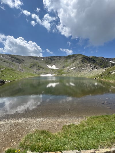 A serene, sunlit mountain lake reflects the blue sky and fluffy white clouds. The water is clear, revealing a rocky bottom near the shore. The lake is surrounded by rolling hills covered in green grass and patches of snow. The barren, rocky slopes of a mountain rise in the background, partially covered by vegetation. The scene conveys a sense of natural tranquility and isolation, typical of a remote alpine environment. There are no people or man-made structures visible in the image. The location is unknown, but the scenery suggests a mountainous region, possibly in the Balkans given the context of Bulgaria.