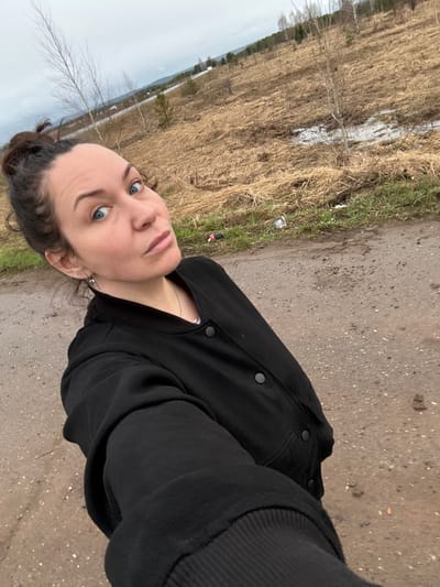 A selfie depicts a woman with light skin, blue eyes, and dark hair pulled back into a bun, wearing a black jacket and necklace. She stands outdoors on a dirt or gravel path. To her left is a field with patches of standing water and bare trees. The sky is overcast. The location is consistent with a rural or semi-rural area. Without any additional context or metadata, it is not possible to confirm or deny the specific location of Povarenki, Russia.
