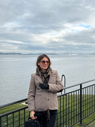 A female subject stands outdoors in Blackness, United Kingdom, positioned in front of a black metal railing overlooking a body of water. She is wearing a brown tweed-patterned blazer with elbow patches, a black and brown patterned scarf, dark trousers, and black gloves. She has light brown hair, wears brown-tinted sunglasses, and a white earbud in her right ear. Her left hand holds a black handbag and rests on the railing, while her right hand is near her chest. She faces towards her left, exhibiting a slight smile. The background features a wide body of water with three bridges visible in the distance: the Forth Bridge, the Forth Road Bridge, and the Queensferry Crossing, beneath an overcast sky with patches of blue. A distant landmass with some structures is visible on the far left horizon.