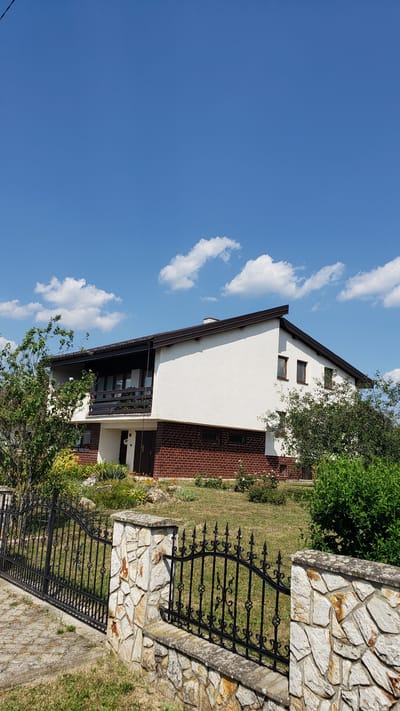 The image shows the side of a white house with a brown roof and brick accents. The house has several windows and a balcony. In front of the house is a black metal fence with ornate designs, which is supported by stone pillars. A stone wall borders the property. The yard is covered in grass and features some trees and plants. The sky is blue with scattered white clouds. The scene appears to be set on a sunny day. The location is Vyšné Nemecké, Slovakia. There is no visible text in the image.