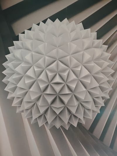 The image shows a white, geometric sphere, resembling a geodesic dome or a spiky orb, suspended in what appears to be an indoor space. The sphere is intricately constructed from numerous triangular facets that create a textured, three-dimensional surface. The background consists of abstract, folded planes in shades of gray and white, suggesting an artistic installation or architectural feature. There are no people visible in the image. The scene is static and appears to be a photograph of a decorative object. This image is not of a specific location in Гарочыцкі сельскі Савет, Belarus, as there is no information to suggest a connection to that place.