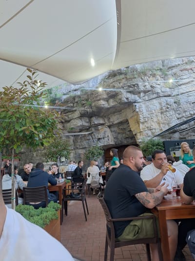 The scene is an outdoor restaurant with patrons seated at wooden tables. The restaurant is constructed at the base of a rock cliff face, with greenery interspersed. People are eating and drinking. The location is Milanovo, Bulgaria.